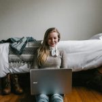 woman sitting beside a bed while using a laptop